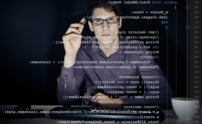 young man with glasses sitting in front of his computer, programming. the code he is working on can be seen through the screen.